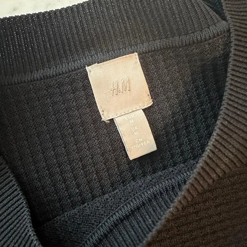 Säljer en stilren svart tröja från H&M med dekorativa knappar på axeln. Tröjan har en ribbad struktur och trekvarts ärmar, perfekt för kyligare dagar. . Puserot.