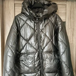 Jättesnygg grön quiltad vinterjacka! - Snygg grön quiltad jacka från Mohito med luva och dragkedja. Jackan har en glansig yta och stora fickor framtill. Perfekt för kyligare dagar.