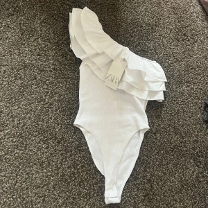Vit oneshoulder body från Zara - Snygg vit oneshoulder body från Zara med volangdetaljer. Perfekt för en stilren look med en touch av elegans. Passar utmärkt till både jeans och kjol.