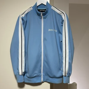 Blå träningsjacka från Palm Angels - Snygg ljusblå träningsjacka från Palm Angels med vita ränder längs ärmarna och dragkedja framtill. Jackan har en sportig look med hög krage och ribbade muddar.