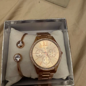 Guldfärgad klocka från Regal - Elegant guldfärgad klocka från Regal med glittrande stenar runt urtavlan. Klockan har tre små urtavlor som och romerska siffror. Armbandet är i metall och matchar med ett stilrent armband med två  stenar.klockan behöver byta batteri 