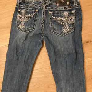 Blå jeans från Miss Me - Snygga blå jeans från Miss Me med broderade detaljer och pärlor på bakfickorna. Jeansen har en skinny passform och låg midja, perfekt för en trendig look. De är tillverkade i denim med en klassisk femficksdesign och knappar i silverfärg.