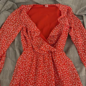 Röd blommig klänning - Säljer en röd blommig klänning med volangdetaljer och omlott-design. Klänningen har långa ärmar och en v-ringad hals. Perfekt för sommaren.