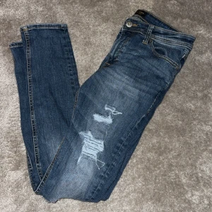 Jack & Jones jeans - Snygga blå jeans med slitningar på benen för en trendig look. De har en klassisk femficksdesign och knappgylf. Perfekta för en avslappnad stil.
