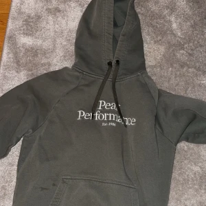 Grå hoodie från Peak Performance - Säljer en grå hoodie från Peak Performance med tryck framtill. Den har en klassisk design med huva och dragsko samt en stor ficka på magen. Perfekt för en avslappnad stil.