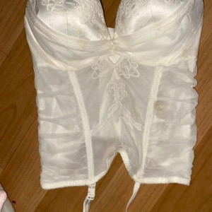 Vit korsett med spetsdetaljer - Elegant vit korsett med vackra spetsdetaljer och vadderade kupor. Den har en smickrande passform och stängs med hyskor i ryggen. Perfekt för speciella tillfällen. 80c 