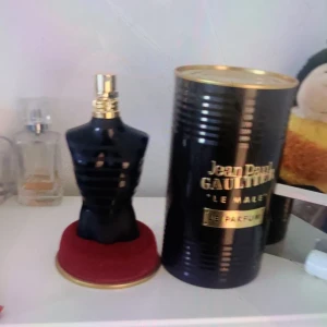 Le Male Le Parfum från Jean Paul Gaultier - Säljer en elegant parfymflaska formad som en svart manlig torso med gulddetaljer från Jean Paul Gaultier. Flaskan står på en röd bas och kommer med en svart och guldig förpackning. Perfekt för den som vill ha en stilren och maskulin doft.