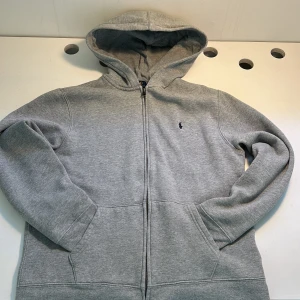 Grå hoodie från Polo Ralph Lauren - Säljer en klassisk grå hoodie från Polo Ralph Lauren med dragkedja och broderad logga på bröstet. Tröjan har en bekväm passform med långa ärmar och en praktisk huva. Perfekt för en avslappnad stil.