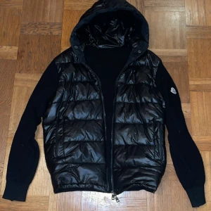 Svart dunväst med huva från Moncler - Snygg svart dunväst från Moncler med dragkedja. Perfekt för kyliga dagar och ger en stilren look. Västen har Moncler-logga på ärmen. Cardiganen är använd 3 gånger och säljer för att spara till bilkörkort.