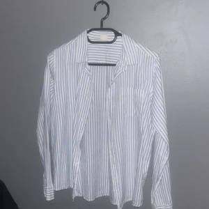 Vit och blå randig skjorta - Snygg vit skjorta med blå ränder. Skjortan har långa ärmar och en klassisk krage. Perfekt för en stilren look.