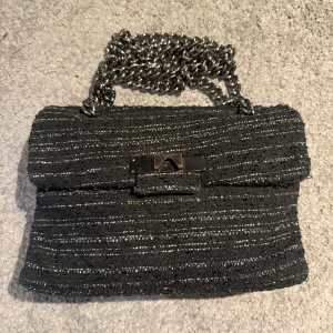 Svart glittrig axelväska från Kurt Geiger - Snygg svart axelväska från Kurt Geiger med glittrigt mönster och kedjehandtag. Väskan har en stilren design med ett metallspänne framtill. Perfekt för att ge en touch av glamour till din outfit.