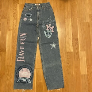 Jeans med tryck från Jennyfer - Coola jeans från Jennyfer med tryck och broderade stjärnor. De har en straight passform och är dekorerade med texten 'Have Fun' och 'East Coast NY'. Perfekta för en avslappnad stil.
