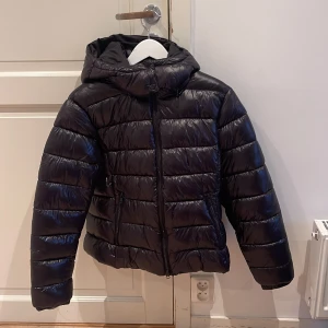 Svart pufferjacka med huva - Snygg svart pufferjacka med huva och dragkedja. Jackan har en glansig yta och är perfekt för kyligare dagar. Den har långa ärmar och en stilren design som passar till många olika outfits.