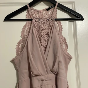 Ljusrosa klänning med spetsdetaljer - Elegant ljusrosa klänning med spetsdetaljer och omlott-design. Klänningen har en vacker spetsrygg och en subtil slits framtill. Perfekt för speciella tillfällen. Finns dessutom ett band som går att knyta till en vacker rosett! 