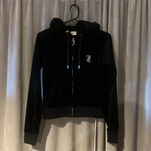 Svart velourjacka från Juicy Couture XS - Snygg svart velourjacka från Juicy Couture med dragkedja och huva. Jackan har glittrande detaljer med logotypen på bröstet och texten 'Juicy' på ryggen. Använd en gång, som ny!!