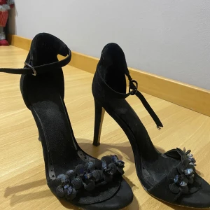 Svarta klackskor med blommor - Eleganta svarta klackskor med blommiga detaljer framtill. Skorna har en smal rem runt ankeln och en hög klack som ger en stilren look. Perfekta för en kväll ute eller en speciell tillställning. Priset kan diskutera
