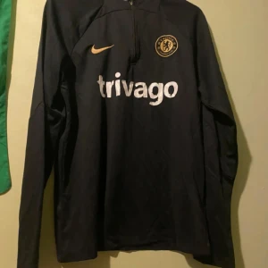 Svart långärmad fotbollströja från Nike - Snygg svart långärmad fotbollströja från Nike med Chelsea FC-logga och trivago-tryck på bröstet. Perfekt för fans som vill visa sitt stöd. Tröjan har en dragkedja vid halsen för justerbar komfort.