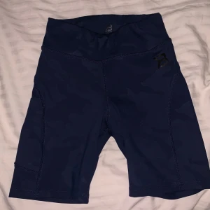 Mörkblå träningsshorts från Better Bodies - Säljer ett par mörkblå träningsshorts från Better Bodies. De är högmidjade och har en bekväm passform, perfekta för gymmet eller löprundan. Materialet är stretchigt och andas bra. Har en camouflage design, se sisa bild.