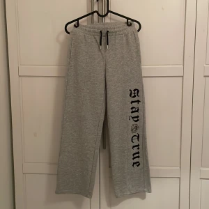 Grå mjukisbyxor med tryck - Snygga grå mjukisbyxor med texten 'Stay True' tryckt längs benet. Byxorna har en elastisk midja med snörning för justerbar passform. Perfekta för en avslappnad stil.