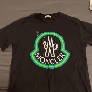 Svart t-shirt från Moncler - Snygg svart t-shirt från Moncler med stort tryck i grönt och blått på framsidan. T-shirten har korta ärmar och en klassisk rund halsringning. Perfekt för en stilren look.