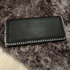 Svart plånbok från Gina Tricot - Snygg svart plånbok från Gina Tricot med kedjedetaljer längs kanterna. Den har en praktisk dragkedja inuti för säker förvaring av dina tillhörigheter. Perfekt för att hålla ordning på kort och kontanter.