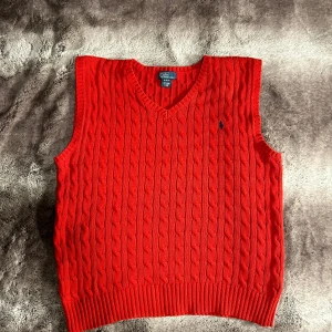 Polo Ralph Lauren - Säljer en snygg röd stickad väst från Polo Ralph Lauren med klassiskt kabelstickat mönster och v-ringning. Perfekt för att lägga till en färgklick i din outfit. Den har en liten broderad logga på bröstet.