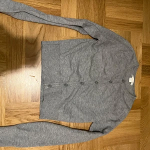 Grå kofta med knappar - Säljer en stilren grå kofta med knappar framtill. Den har långa ärmar och ribbade muddar vid ärmslut och nederkant. Perfekt för lager-på-lager under kyligare dagar.