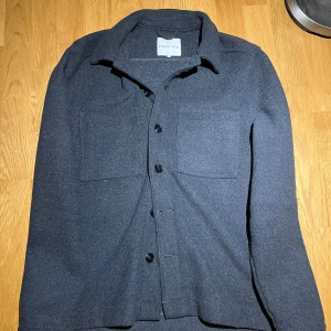 Mörkgrå overshirt från Studio Total - Snygg mörkgrå overshirt från Studio Total med knappar framtill och två bröstfickor. Perfekt för lager-på-lager-stil. Tillverkad i ett mjukt material som ger en bekväm passform. För liten för mig därför säljer, knappt använd