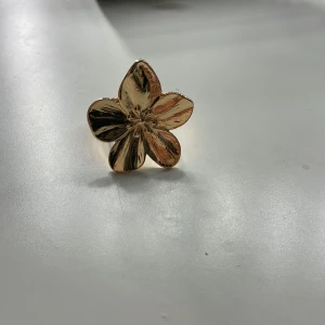 Guldfärgad blomformad ring - Elegant guldfärgad ring i form av en blomma med fem kronblad. Ringen har en justerbar design för att passa olika storlekar. Perfekt för att ge en touch av glamour till din outfit.