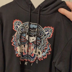 Svart hoodie med tigerbroderi från Kenzo - Snygg svart hoodie från Kenzo med ett stort, färgglatt tigerbroderi på framsidan. Hoodien har en justerbar dragsko i huvan och en klassisk passform. Perfekt för en cool och avslappnad stil.