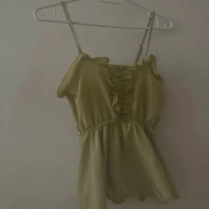 Olivgrön topp med volangdetaljer - Snygg olivgrön topp med smala axelband och volangdetaljer framtill. Perfekt för varma sommardagar eller en avslappnad kväll. Den har en luftig passform och är tillverkad i ett lätt material. Köpt i Sydkorea