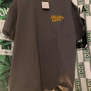 Grå t-shirt från Gallery Dept. - Snygg grå t-shirt från Gallery Dept. med gul text på bröstet och ryggen. Perfekt för en avslappnad stil.