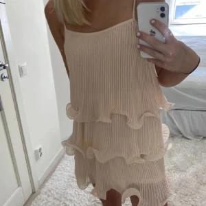 Beige plisserad klänning - Snygg beige klänning med plisserade detaljer och volanger. Perfekt för en somrig look. Klänningen har en kort längd och är ärmlös, vilket gör den luftig och bekväm. Den är sydd innuti men går att ta bort💞
