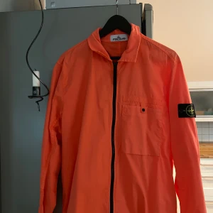 Orange jacka från Stone Island - Snygg orange jacka från Stone Island med dragkedja och en bröstficka. Jackan har en ikonisk logga på ärmen och är perfekt för en stilren look.