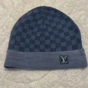 Blå rutig mössa från Louis Vuitton - Snygg blå mössa från Louis Vuitton med ett klassiskt rutigt mönster. Mössan har en uppvikt kant med LV-logotypen framtill. Perfekt för att hålla stilen i kallare väder.