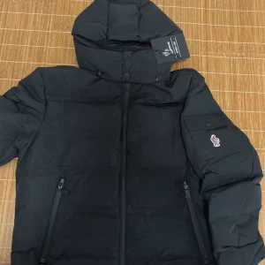 Moncler Grenoble  - Snygg svart dunjacka från Moncler med huva och dragkedja. Jackan har praktiska fickor och en stilren design, perfekt för kyliga dagar. OBS kan ej garantera att jackan är helt äkta!!