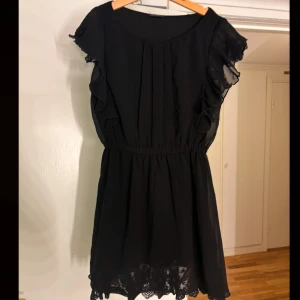 Svart klänning med spetsdetaljer - Elegant svart klänning med spetsdetaljer vid fållen och volangärmar. Perfekt för en stilren look med en touch av romantik. Den har en smickrande passform med elastisk midja.