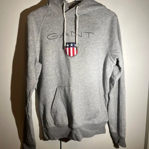 Grå hoodie från GANT - Säljer en stilren grå hoodie från GANT med broderad logga och ett amerikanskt inspirerat emblem på bröstet. Tröjan har en justerbar huva med snören och en praktisk magficka. Perfekt för en avslappnad look! 