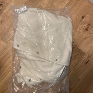 Vit teddyjacka med tryckknappar - Mysig vit teddyjacka med tryckknappar framtill och två stora fickor. Perfekt för kyliga dagar när du vill hålla dig varm och bekväm. Jackan har en enkel och stilren design som passar till det mesta.