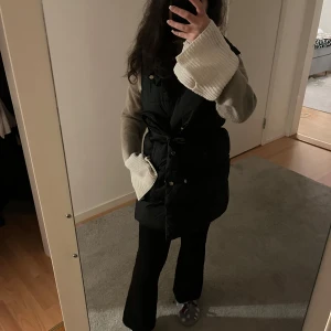 Svart lång pufferväst - Säljer en svart lång pufferväst med knappar och bälte i midjan. Jackan har en stilren design och är perfekt för kyligare dagar. Den är lätt att matcha med olika outfits och ger en trendig look. Den är ifrån Giorgia fashion och är i storlek S!