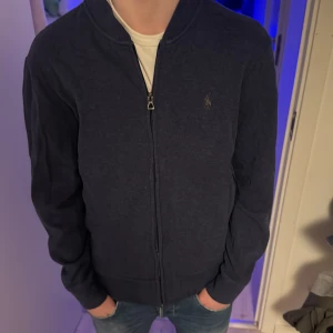 Mörkblå zip-tröja från Polo Ralph Lauren - Säljer en stilren mörkblå zip-tröja från Polo Ralph Lauren. Tröjan har en dragkedja framtill och en liten broderad logotyp på bröstet. Perfekt för en avslappnad och snygg look.