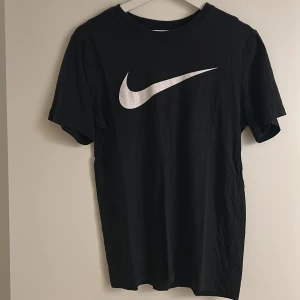 Svart t-shirt från Nike - Säljer en klassisk svart t-shirt från Nike med den ikoniska vita swoosh-loggan på bröstet. Perfekt för en sportig look eller avslappnad stil. Tillverkad i mjukt material för extra komfort.