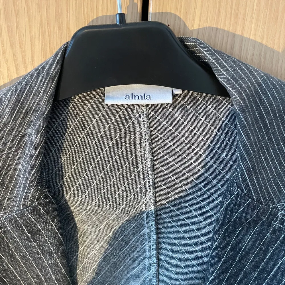 Snygg grå kavaj från Almia med smala vita ränder och knappar framtill. Kavajen har en klassisk krage och praktiska fickor. Perfekt för en stilren look.. Takit.