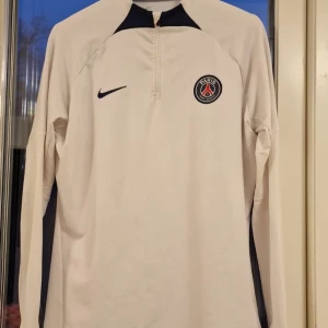 Vit långärmad tröja från Nike med PSG-logga - Säljer en vit långärmad tröja från Nike med PSG-logga på bröstet. Tröjan har en dragkedja vid halsen och är tillverkad i Dri-FIT-material för optimal komfort. Perfekt för fotbollsfans som vill ha en stilren look.