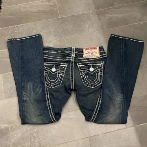 Mörkblå jeans från True Religion - Snygga mörkblå jeans från True Religion med kontrasterande vita sömmar och klassisk femficksdesign. Jeansen har en låg midja perfekt för en avslappnad stil. Märkeslogga på bakfickan och knappar i silverfärg. Finns en liten fläck som säkert går bort med medel.  Några är lånade bilder från förra ägaren, säljer då de är för korta för mig.
