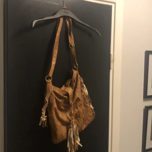 Brun axelväska med fransar - Snygg brun axelväska  med fransar. Väskan har justerbar axelrem och dekorativa detaljer som ger en bohemisk känsla. Perfekt för att ge din outfit en unik touch.