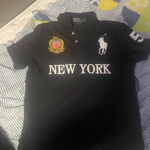  Svart pikétröja från Ralph Lauren / chief keef - Snygg svart pikétröja från Ralph Lauren med broderad logga och texten 'New York' på framsidan. Tröjan har en stor siffra '5' på ryggen och en krage med knappar. Perfekt för en sportig look!