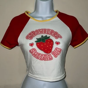 Vit och röd croppad t-shirt med jordgubbstryck - Säljer en söt croppad t-shirt i vitt och rött med texten 'Strawberry Sweetie Pie' och ett stort jordgubbstryck på framsidan. T-shirten har korta ärmar och en gul kant vid halsen. Köpte dem från Shein 50-60kr 