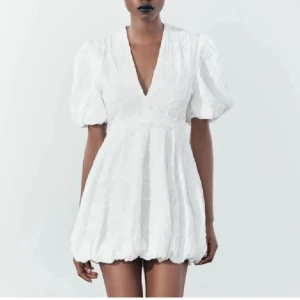 Vit kortklänning med puffärmar - Trendig vit kortklänning med djup v-ringning och puffiga kortärmar. Klänningen har en unik struktur och en ballongformad nederdel som ger en lekfull look.