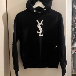 Svart hoodie och byxor med YSL-logga - Snygg svart hoodie och matchande byxor med vit YSL-logga. Hoodien har en klassisk design med huva och långa ärmar. Byxorna har dragsko i midjan för justerbar passform. Perfekt för en stilren och bekväm look.